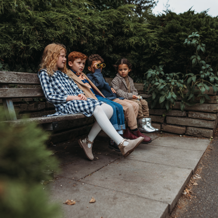 Four children sitting together on a wooden park bench, surrounded by green bushes and trees, with a paved path and stone wall behind them.