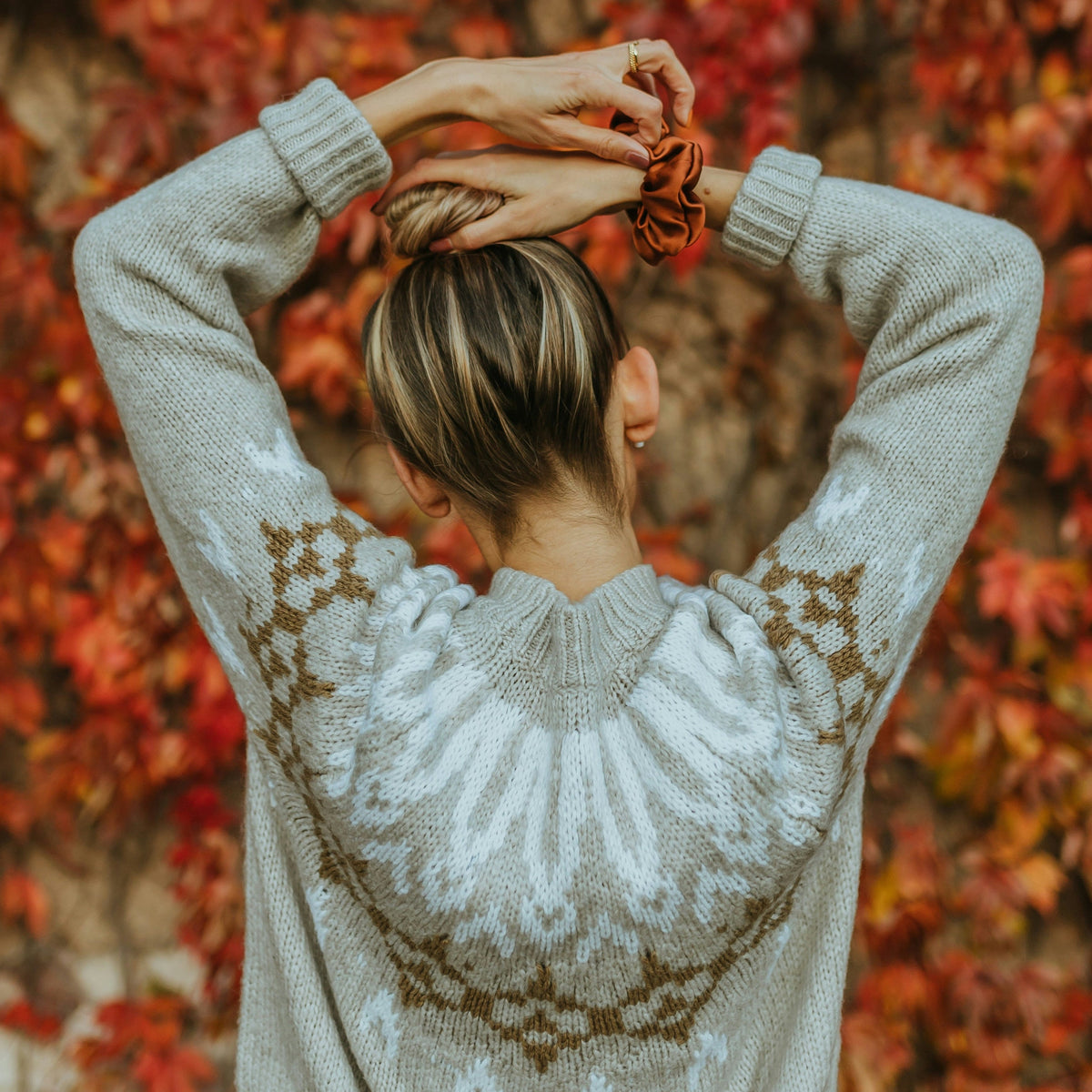 A woman viewed from behind wearing a light gray knit sweater with a brown Fair Isle pattern, standing in front of a dense background of red autumn leaves.