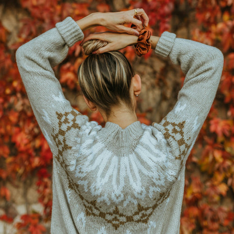 A woman viewed from behind wearing a light gray knit sweater with a brown Fair Isle pattern, standing in front of a dense background of red autumn leaves.