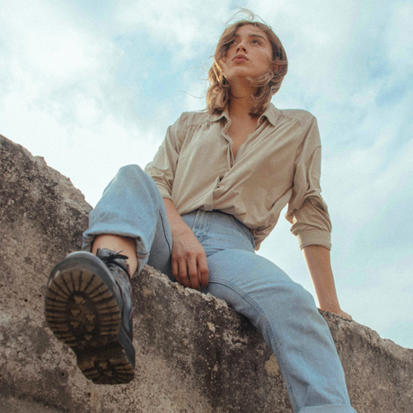 A woman sits on a rough stone ledge outdoors under a cloudy sky, wearing a beige button-up shirt, light blue jeans, and a dark lace-up boot.