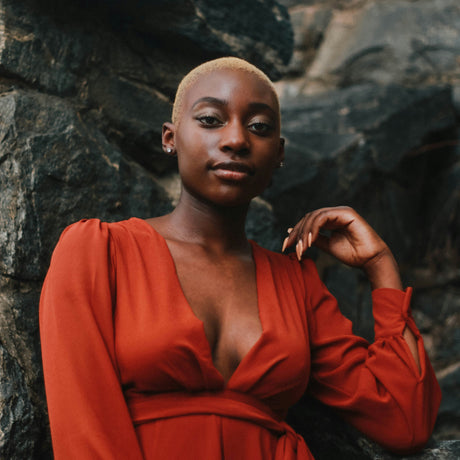 A woman with a shaved head wearing a rust-orange long-sleeve wrap dress, posing against a dark, textured rock wall.