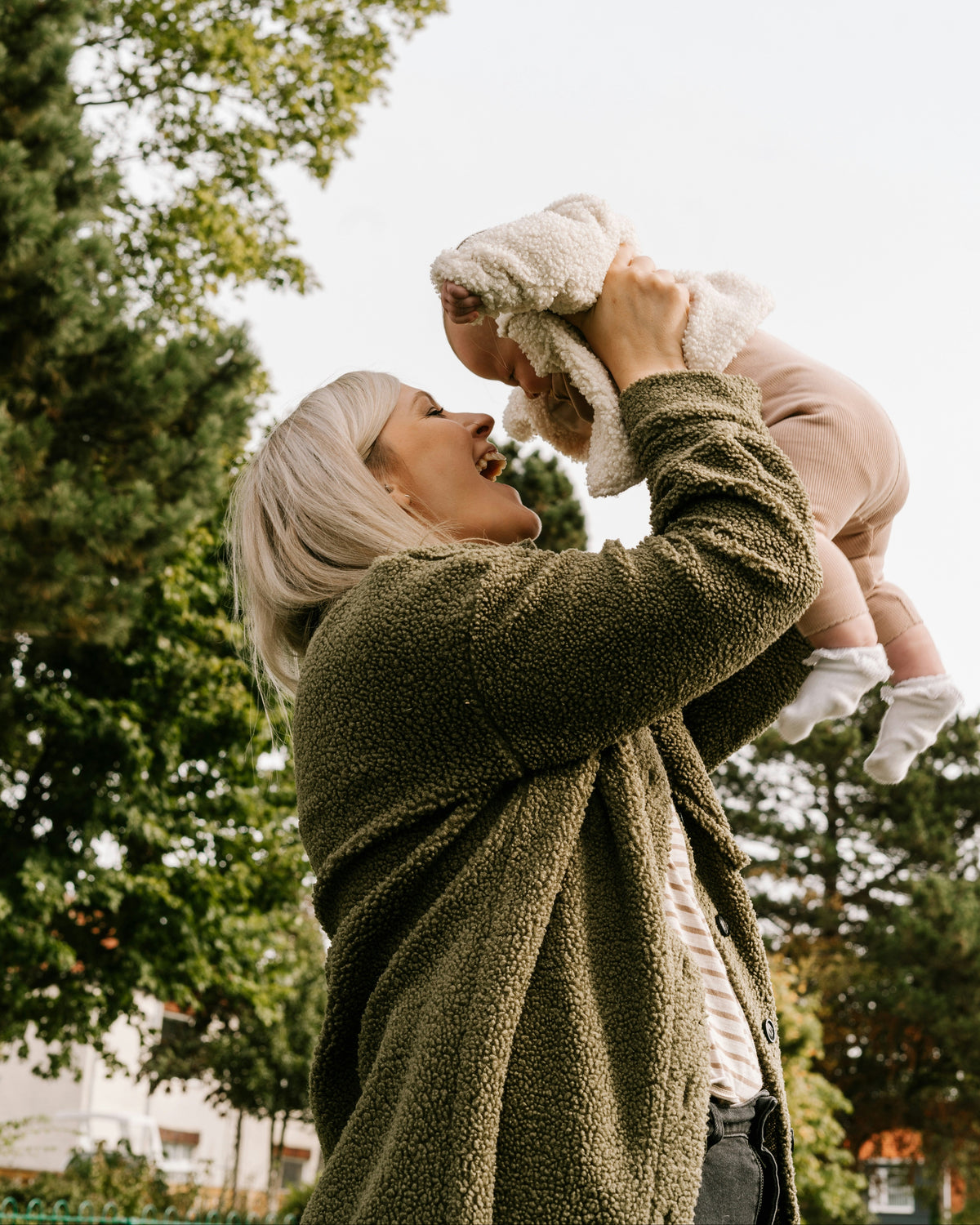 A woman in an olive green fleece jacket holds a baby up in the air outdoors, smiling and laughing. The baby is wrapped in a cream blanket and wearing light pink clothing.